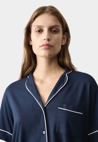 Haut de pyjama à manches courtes bleu marine en tissu doux, doté d'un passepoil blanc le long du col et d'une poche poitrine avec une finition assortie.