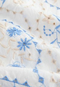 Tissu blanc avec broderie florale bleue et beige comprenant des fleurs, des tiges et de petits œillets dans un motif délicat.