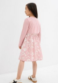 Rosa Kleid mit langen Ärmeln, das ein Blumenmuster am Rock und eine geraffte Taille aufweist. Tragen mit beige-weißen Turnschuhen.