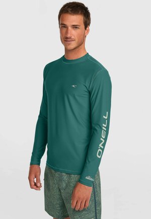 Man met een tealkleurig O'Neill rashguard met lange mouwen en groene zwembroek met bloemenpatroon, staand tegen een effen lichte achtergrond.