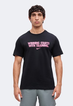 Jeune homme aux cheveux bouclés foncés portant un t-shirt noir avec le texte rose « Winning starts with training » et un short gris, debout devant un fond uni.