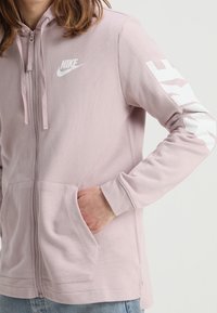 Světle růžová mikina na zip s přední kapsou, bílým logem na hrudi a bílými detaily na rukávech. Měkká látka, volný střih.