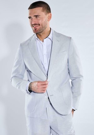 Mann trägt hellgrauen Anzug und weißes Hemd, knöpft die Jacke zu, lächelt, Hand in der Tasche, vor schlichtem hellem Hintergrund.