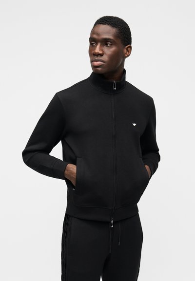 Svart hettejakke med glidelås, høyt krage, sidelommer og ribbestrikkede mansjetter. Brodert logo på brystet. Myk, glatt stofftekstur.