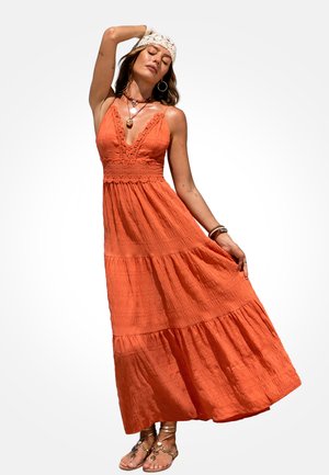Frau in orangefarbenem ärmellosem Maxikleid und gehäkeltem Stirnband posiert mit einem erhobenen Arm und Sandalen an den Füßen vor einfachem Hintergrund.
