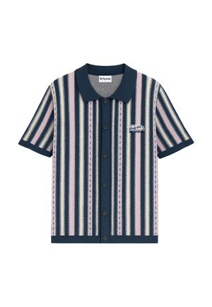 Man with curly hair wearing matching short-sleeve button shirt and pants with vertical navy, pink, and white stripes, hands in pockets.