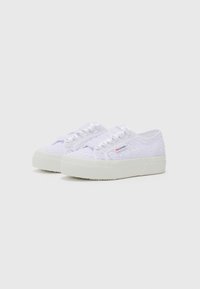 White lace sneakers with textured fabric, flat white rubber sole, and satin ribbon laces. Features floral lace pattern and subtle branding.