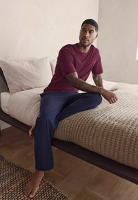 Bordowa koszulka z krótkim rękawem z kieszenią, zestawiona z granatowymi spodniami. Osoba siedzi na teksturowanym, dzianym kocu na łóżku z neutralną pościelą.
