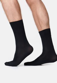 Zwei barfuß Füße in schwarzen Socken, ein Fuß leicht angehoben. Der Hintergrund ist schlicht weiß, was die Socken und Beine betont.