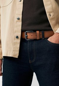 Belt din piele maro cu o cataramă din metal negru mat, purtat peste blugi denim de culoare albastră închisă și asortat cu o cămașă bej cu nasturi.