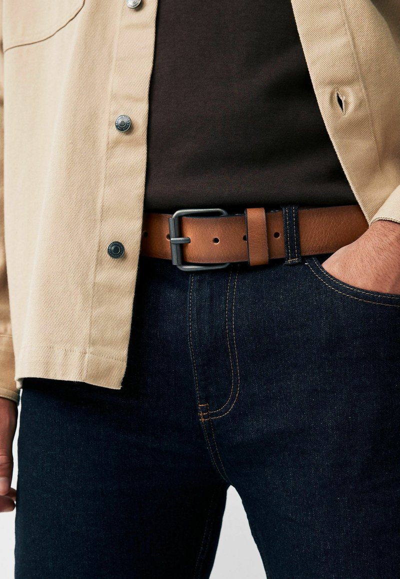 Belt din piele maro cu o cataramă din metal negru mat, purtat peste blugi denim de culoare albastră închisă și asortat cu o cămașă bej cu nasturi.