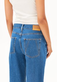 A model wears high-waisted blue jeans with a brown label, showcasing the back view and pockets against a plain white background.