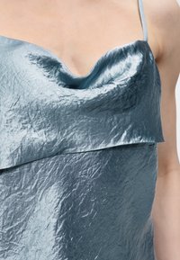 Γυναίκα που φοράει ένα ασημί-μπλε σατινέ καμιολέ με λαιμόκοψη Cowl, λεπτές τιράντες στον ώμο και υφή στο ύφασμα.