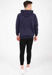 Marinblå hoodie med dragsko i huvan, ribbade manschetter och nederkant. Kombinerad med slim-fit svarta byxor och vita sneakers, sedd bakifrån.