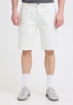 Mand iført hvide shorts til knæet, hvide crew-sokker og lysegrå sneakers, stående mod en ensfarvet baggrund.