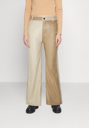 Zweifarbige beige Hose mit weitem Bein, getragen mit schwarzen Schuhen und einem langärmligen Pfirsichton-Oberteil, gezeigt vom mittleren Oberkörper bis zu den Füßen vor einem schlichten Hintergrund.