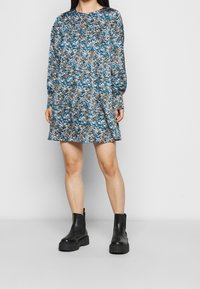 Květinové dlouhé šaty s dlouhým rukávem v modré, hnědé a bílé barvě; volný střih s kulatým výstřihem. Nosené s černými chunky kotníkovými botami.