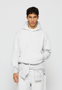 Hellgrauer Hoodie aus einem weichen Stoff, mit einer Kängurutasche, einer Kapuze mit Zugband und gerippten Bündchen. Getragen mit passenden Hosen.
