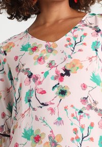 Blusa a fantasia floreale con scollatura a V. Presenta fiori colorati in rosa, verde e blu su uno sfondo rosa chiaro. Tessuto morbido e fluido.