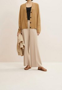 Cárdigan de punto beige con botones negros, llevado sobre una blusa negra, combinado con pantalones anchos beige y sandalias deslizantes marrones.