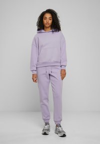 Ensemble en hoodie et jogging lavande avec une poche avant, des poignets côtelés et une taille à cordon. Associé à des chaussures de sport grises.