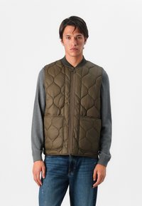 Kurtka pikowana w oliwkowym kolorze z zamkiem z przodu, okrągłym kołnierzem i dwiema dużymi kieszeniami. Nosi się ją na szarym longsleeve'ie, o dopasowanym kroju.