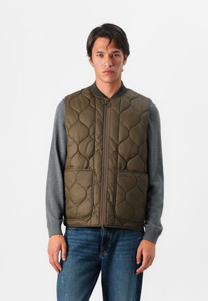 Quilted olive green vest with front zipper, rounded collar, and two large pockets. Worn over a grey long-sleeve shirt, slim fit design.