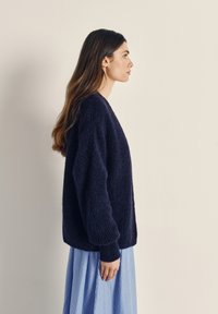 Navy blue oversized sweater with a textured knit pattern, featuring a deep V-neck and cuffed sleeves, worn over a light striped skirt.
