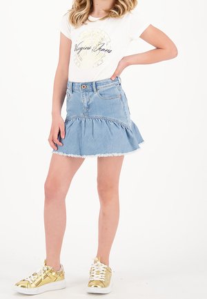 Mädchen trägt weißes T-Shirt mit Logo, hellblauen Jeans-Rüschrock und goldene Metallic-Sneaker, steht mit einer Hand in der Hüfte.