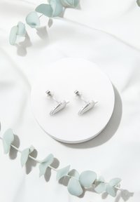 Silverfärgade, morotsformade cufflinks med texturerad detaljering, graciöst placerade på en rund vit yta, omgivna av mjukt grönt på en ljus tygbakgrund.