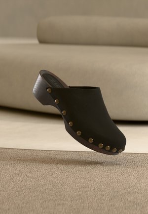 Sabot en bois noir avec des clous en laiton le long du bord de la semelle, flottant au-dessus d'un tapis beige dans une pièce aux tons neutres.