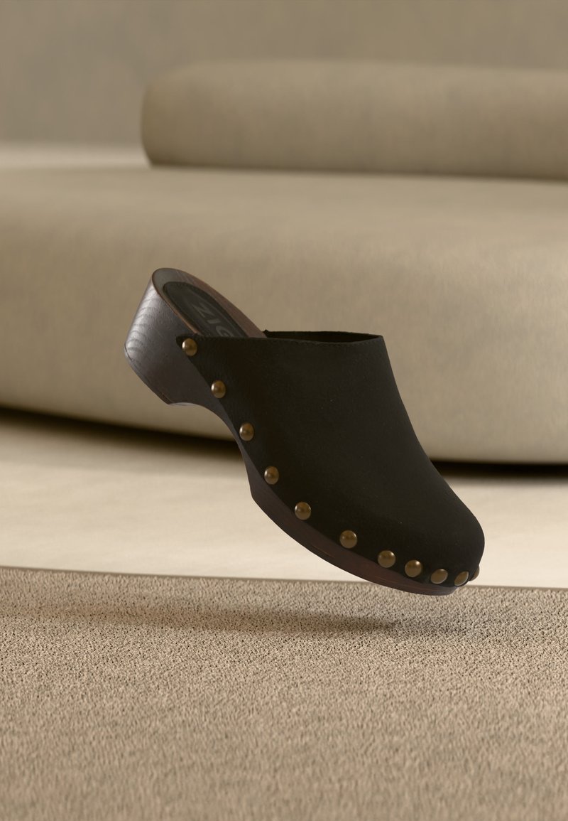 Sabot en bois noir avec des clous en laiton le long du bord de la semelle, flottant au-dessus d'un tapis beige dans une pièce aux tons neutres.