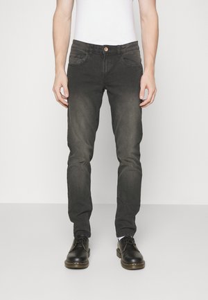 Homme portant un jean noir délavé slim-fit et des chaussures noires à lacets, debout devant un fond blanc uni.