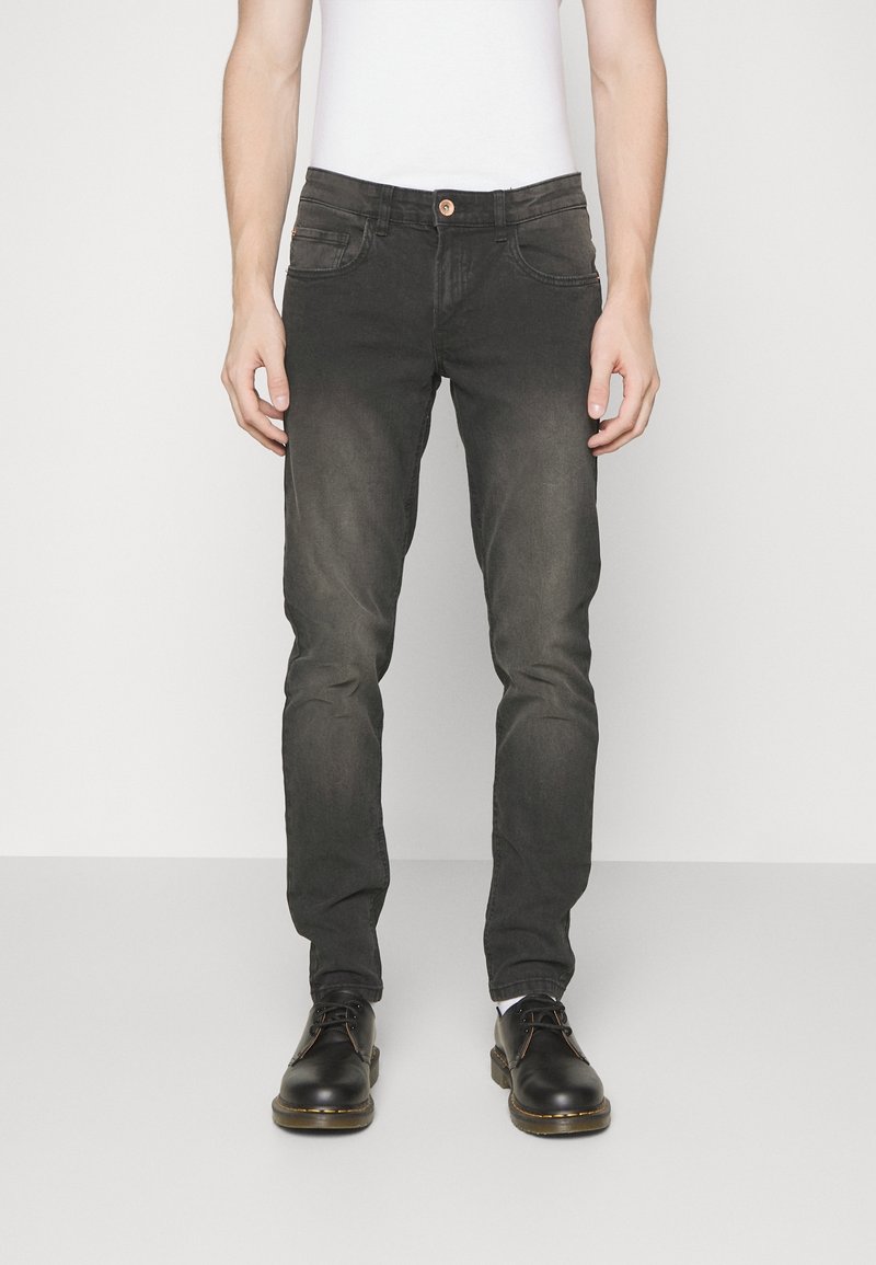 Homme portant un jean noir délavé slim-fit et des chaussures noires à lacets, debout devant un fond blanc uni.