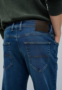 Tamno plave traperice s stražnjim džepovima i kontrastnim narančastim šavovima. Pojas sadrži smeđu kožnu etiketu s utisnutim natpisom "SALSA JEANS".