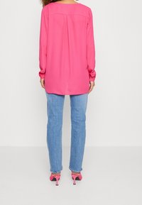 Blouse à manches longues, rose vif, avec un col arrondi et un détail plissé au dos, associée à un jean bleu clair et des chaussures à talons roses.