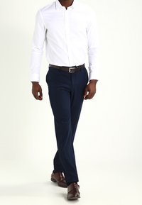 Camisa blanca de manga larga, pantalones azul marino, zapatos de cuero marrón. La camisa tiene un cuello estándar y cierre de botones; los pantalones tienen un ajuste slim.
