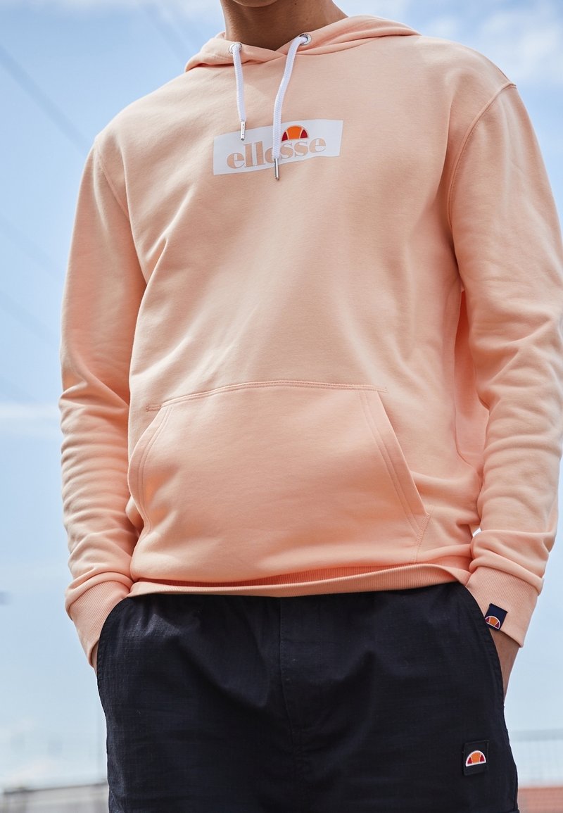 Personne portant un sweat à capuche couleur pêche clair avec cordons blancs et un pantalon noir, tous deux arborant le logo de la marque ellesse.