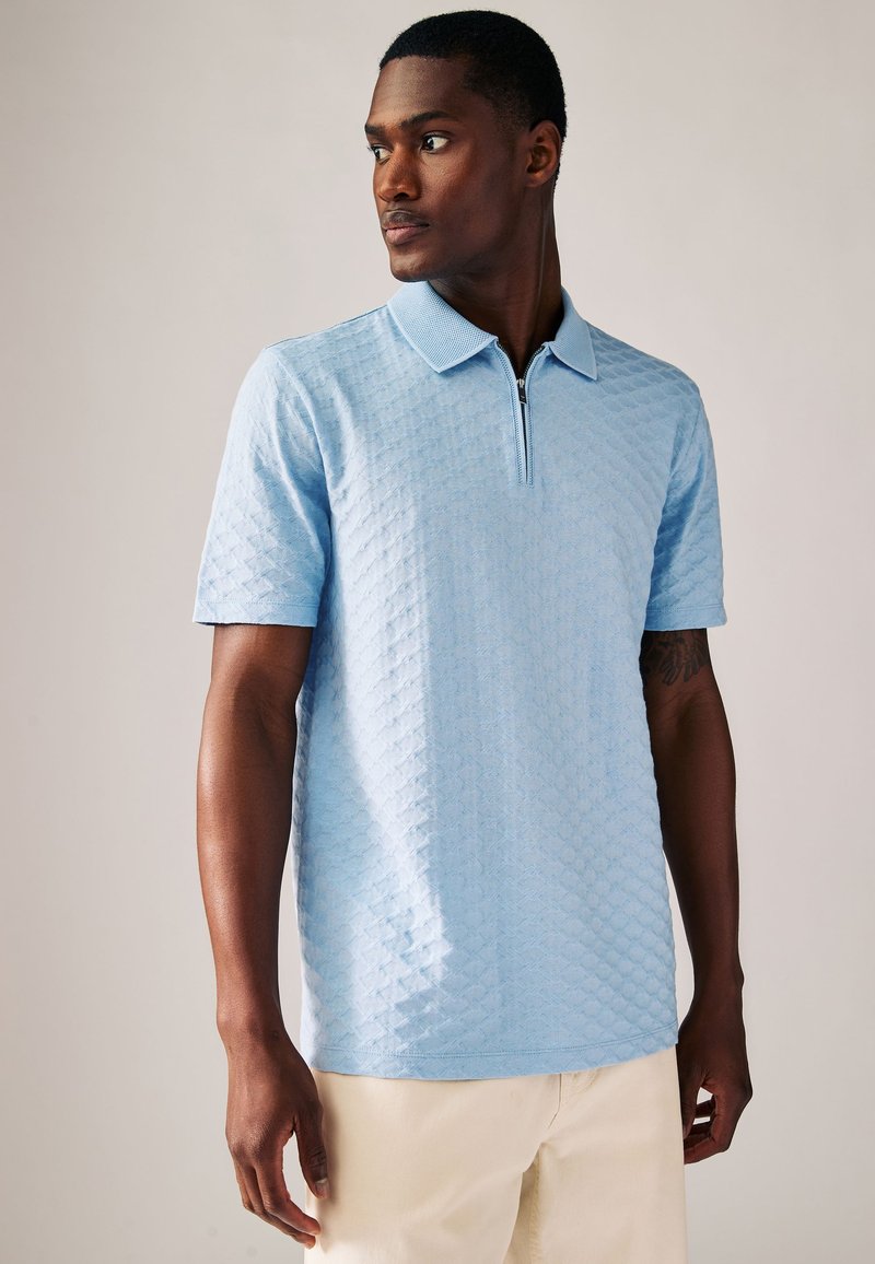 Světle modré polo tričko s límečkem na zip, strukturovaným diamantovým vzorem, krátkým rukávem a volným střihem.