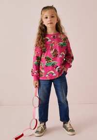 Felpa rosa luminoso con motivi colorati di auto, arcobaleni e alberi. Indossata con jeans blu e sneaker argento glitterati. Bambino tiene anelli di carta.