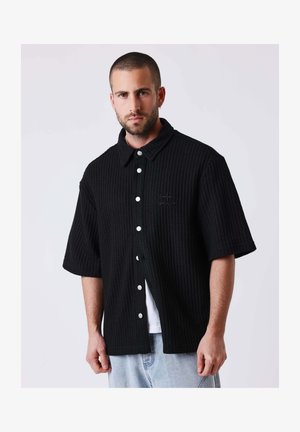 Homme aux cheveux courts et à la barbe portant une chemise noire texturée à manches courtes boutonnée sur un débardeur blanc et un jean bleu clair.