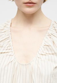 Κοντινό πλάνο του λαιμού και της κλείδας ενός ατόμου που φοράει κρεμ μπλούζα με λεπτές κάθετες ρίγες και βολάν στους ώμους.