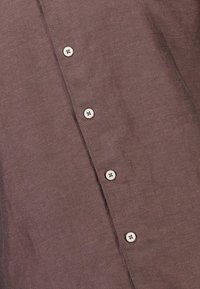 Bruine stof met een rij van vier witte knopen die verticaal zijn genaaid langs een fijn gestikte naad.