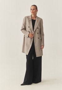 Beige kahekordne villane jope, mustad laiad püksid, mustad terava ninaga jalanõud ja kuldsed erksad kõrvarõngad kandv naine neutraalses taustas.
