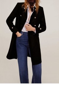 Abrigo negro de doble botonadura con botones plateados, combinado con una camisa blanca y jeans azules. Presenta un corte largo y una textura suave.