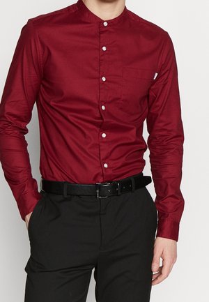 Rotes Baumwollhemd mit Stehkragen, Vorder-pocket und weißen Knöpfen, kombiniert mit schwarzen Hosen und einem schwarzen Gürtel. Glatte Textur.