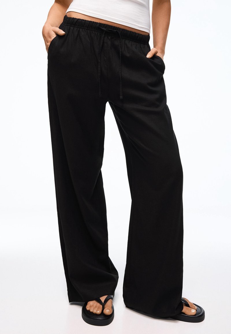 Personne portant un pantalon noir ample avec une taille à cordon, mains dans les poches, associé à des tongs noires et un top court blanc.