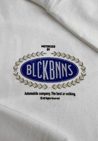 Witte stof met een blauw ovaal logo met "BLCKBNNS," omringd door een lauwerkrans. Onder de tekst staat "Autobedrijf. Het beste of niets."