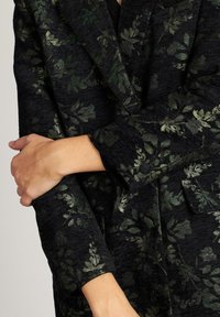 Chaqueta negra con patrón floral en verde oscuro, con puños abotonados y bolsillo con solapa, llevada por una persona con los brazos cruzados.