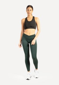 Zwart sportbeha met een aangesloten ontwerp, gecombineerd met donkergroene leggings met hoge taille. Model draagt witte sportschoenen.